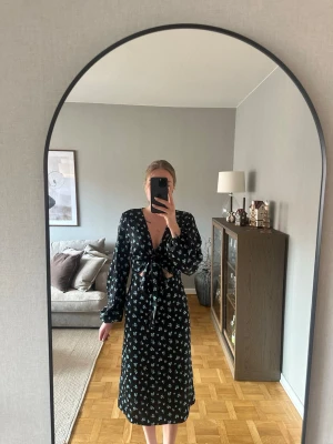 Svart blommig klänning från GinaTricot  - Snygg svart klänning från GinaTricot med små blommor i  blått. Klänningen har långa små ballong ärmar,             v-ringning och knyt framtill. Midilängd. Använda ett fåtal gånger. 