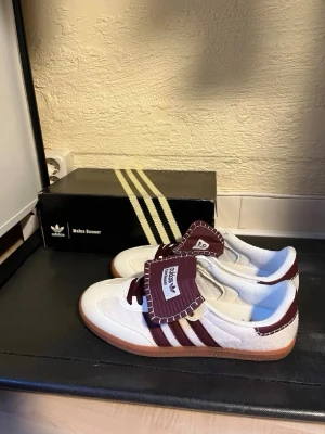 Adidas Wales Bonner Samba vinröd/vit - Låda och kvitto finns! Säljer ett par Adidas Wales Bonner Samba sneakers i vitt och vinrött med klassiska tre ränder och gummisula, de är lite använda och har små märken. Skorna har unika detaljer som kontrastsömmar och extra stor plös med Wales Bonner-logga. Materialet är en mix av läder och mocka för en retrovibe. 