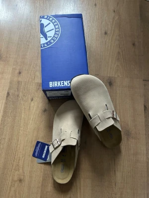 Birkenstock Boston tofflor - Säljer ett par beige Birkenstock Boston tofflor i storlek 45. De är tillverkade i mjukt mockamaterial med justerbar rem och metallspänne. Klassisk design med bekväm innersula och stilren look, perfekt för dig som gillar minimalistiska skor.