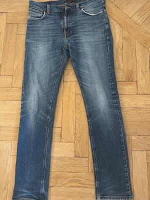 Blå jeans från Nudie Jeans - Säljer ett par klassiska blå jeans från Nudie Jeans med snyggt slitna detaljer och kontrastsömmar. En av de främre fickorna har hål i sig, raka ben och cool brodyr på bakfickorna. Materialet är mjukt denim som sitter skönt och ser riktigt bra ut. Storlek w32 L34
