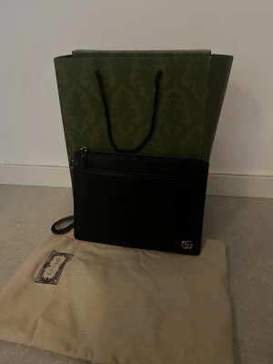 Svart kuvertväska från Gucci - Snygg svart kuvertväska från Gucci i slätt läder med guldfärgad GG-logga i hörnet. Väskan har dragkedja upptill och en avtagbar handledsrem. Perfekt för att bära det viktigaste och ger en lyxig touch till din outfit. Det som medföljer är kuvertväskan, dustbag, kartong och påse. Kvitto finns inte då jag fick bara detta av köparen innan (hon sa att hon slarvade bort den). Sista bilderna visar serienumret