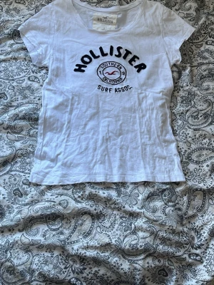 Vit Hollister t-shirt med tryck - Vit t-shirt från Hollister med svart och rött tryck på bröstet där det står 'Hollister Surf Assoc.' och en logga. T-shirten är i barn storlek L men passar XS/S. Klassisk rund hals och korta ärmar. Perfekt för dig som gillar en enkel och stilren look. Passar perfekt till sommaren. Pris kan själv klart diskuteras