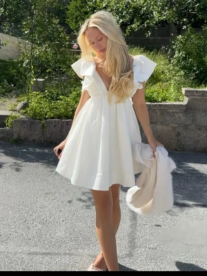 Vit kortklänning med volangärmar - Perfekt till studenten! Helt ny med prislapp! Supersöt vit kortklänning med djup v-ringning och stora volangärmar. Klänningen har en luftig passform och är tillverkad i ett lätt tyg, perfekt för varma dagar. Stilren och trendig design som ger en romantisk vibe.