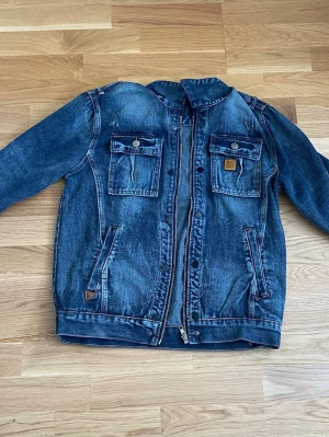 Mörkblå B jeansjacka med dragkedja  - Säljer en snygg mörkblå Burberry jeansjacka med dragkedja och tryckknappar. Jackan har två bröstfickor med lock och knapp, samt två snedställda sidofickor. Klassisk krage och coola slitningar för en trendig look. Perfekt till jeans eller andra byxor.