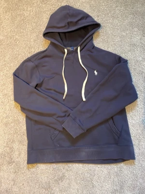 Mörkblå hoodie från Polo Ralph Lauren - Snygg mörkblå hoodie från Polo Ralph Lauren med vit broderad logga på bröstet och vita dragsnören i huvan. Klassisk känguruficka framtill och mjukt material som känns skönt mot huden. Perfekt för en avslappnad och stilren look.