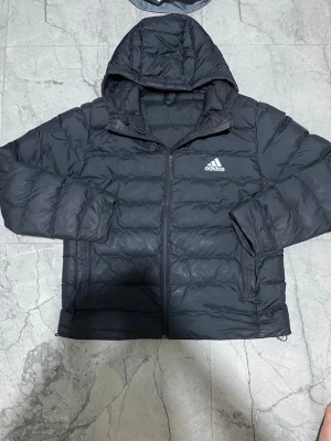 Svart dunjacka med huva från Adidas - Snygg svart dunjacka från Adidas med huva och dragkedja framtill. Jackan har två sidofickor med dragkedja och Adidas-logga på bröstet. Perfekt för kalla vinterdagar och ger en sportig look.