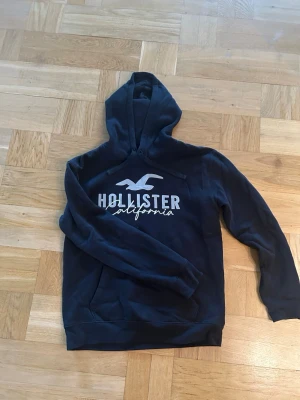 Svart hoodie från Hollister - Svart hoodie från Hollister med stor vit logga och texten 'California' på bröstet. Klassisk huva med dragsko och magficka framtill. Tillverkad i mjukt bomullsmaterial som är skönt att ha på sig.