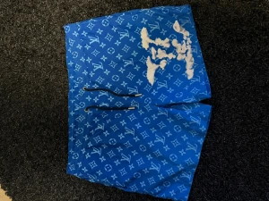 Blå Louis Vuitton badshorts  - Säljer ett par blå shorts från Louis Vuitton med ikoniskt logomönster över hela plagget. Shortsen har snörning i midjan och är tillverkade i ett lätt syntetmaterial, perfekta för sommaren. Snygg och trendig design för dig som vill sticka ut.