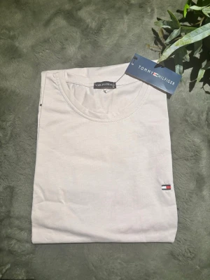Vit t-shirt från Tommy Hilfiger - Snygg vit t-shirt från Tommy Hilfiger med klassisk rund halsringning och liten broderad logga på bröstet. Tillverkad i mjuk bomull som känns skön mot huden. Perfekt basplagg för en clean och stilren look.