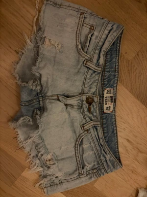 Ljusblå slitna jeansshorts - Säljer ett par ljusblå jeansshorts från Only med råa, fransiga kanter och slitningar för en cool, avslappnad look. Klassisk femficksmodell med knappgylf och detaljer i brunt läder. Perfekta för sommaren och festivaler.