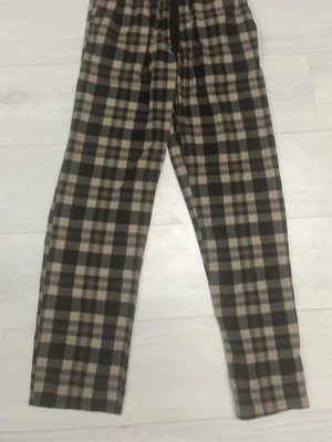 Rutiga kostymbyxor i beige och svart - Snygga kostymbyxor med rutigt mönster i beige, svart och brunt. Byxorna har rak passform och normal midja. Perfekta för dig som vill ha en trendig och stilren look. Materialet känns mjukt och bekvämt.