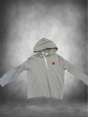 Grå hoodie från Comme des Garçons - Säljer en grå hoodie från Comme des Garçons Play med det ikoniska röda hjärtat på bröstet. Hoodien har huva med vit dragsko och två fickor framtill. Tillverkad i mjukt bomullsmaterial och har en avslappnad passform. Perfekt för en chill och stilren look. Inte orginalsnöret med ingen stor skillnad. 