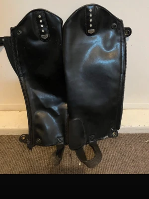 Svarta läderchaps med dragkedja - Säljer ett par svarta chaps i blankt läder med dragkedja längs sidan och tryckknappar upptill. Perfekta för ridning eller motorcykel och har elastiska band under foten för bra passform. Snygg och stilren design.