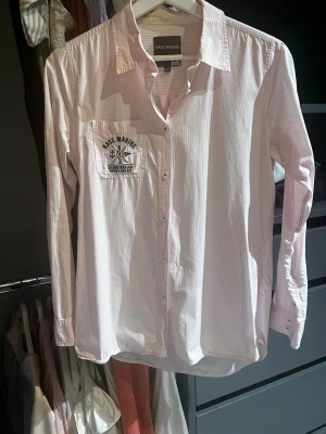 Ljusrosa randig skjorta Race Marine - Säljer en ljusrosa skjorta från Race Marine med tunna vita ränder och tryck på bröstfickan. Skjortan har klassisk krage, knappar framtill och långa ärmar. Perfekt för en avslappnad men ändå stilren look.