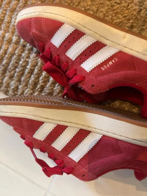 Röda Adidas Campus sneakers mocka - Säljer ett par röda Adidas Campus sneakers med klassiska vita ränder på sidorna och vit sula. Skorna har snörning och den ikoniska Adidas-loggan på sidan. Perfekta för dig som vill ha en retro och sportig look.