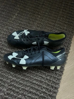 Under Armour svarta fotbollsskor - Snygga svarta fotbollsskor från Under Armour med vita detaljer och stora logotyper på sidan och tån. Skorna har fasta dobbar och snörning för bra passform. Insidan är fodrad i grönt tyg. Perfekta för dig som vill sticka ut på planen.