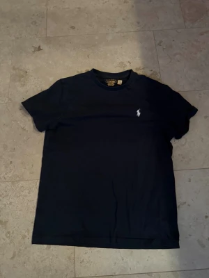 Mörkblå t-shirt från Ralph Lauren - Klassisk mörkblå t-shirt från Ralph Lauren med den ikoniska vita polospelaren broderad på bröstet. T-shirten har rund halsringning och korta ärmar. Tillverkad i mjuk bomull för en skön känsla och enkel stil.