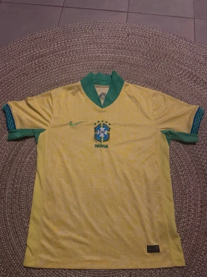 Brasilien matchtröja Nike gul/grön - Säljer en officiell Brasilien matchtröja från Nike i gult med gröna detaljer. Tröjan har korta ärmar, grönt kragparti och blå/gröna muddar. Materialet är lätt och andas, perfekt för fotboll eller träning. Brasilien-emblemet och Nike-loggan finns på bröstet.