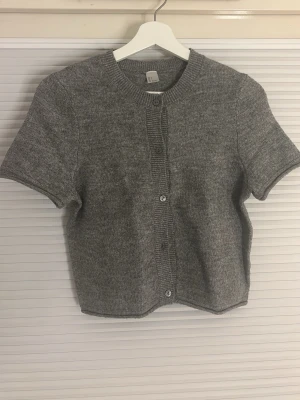 Grå kortärmad kofta med knappar - Kortärmad grå kofta i mjukt stickat material med rund halsringning och knäppning framtill. Klassisk och enkel design som passar till mycket. Ribbstickade detaljer vid ärmslut, hals och knappslå.