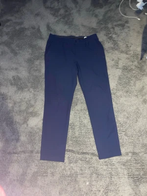 Marinblå golfbyxor från Under Armour - Snygga marinblå golfbyxor från Under Armour med rak passform och klassisk knappstängning. Byxorna har bälteshällor och är tillverkade i ett stretchigt, lätt material som ger skön komfort hela dagen. Perfekta för dig som gillar stilrena och sportiga plagg.