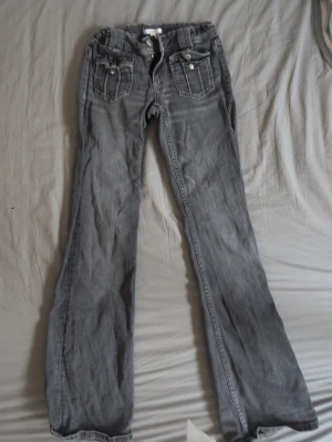 Grå bootcut jeans med fickdetaljer - Säljer ett par grå bootcut jeans med dekorativa fickor framtill och knappar. Jeansen har en klassisk femficksmodell och är tillverkade i denim med lätt tvättad look. Perfekta för dig som gillar en avslappnad men ändå trendig stil. Använda tre gånger