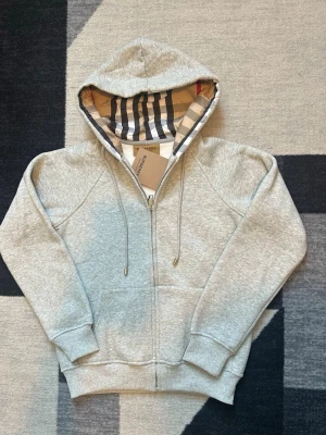 Grå zip hoodie från Burberry - Snygg grå zip hoodie från Burberry med klassiskt rutigt foder i huvan och dragsko. Tillverkad i mjuk bomullsblandning, har fickor fram och ribbade muddar. Perfekt för en chill och stilren look.