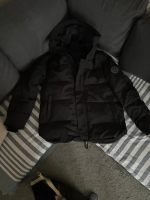 Svart dunjacka från Canada Goose - Snygg svart dunjacka från Canada Goose med huva och diskret logga på ärmen. Jackan har dragkedja och knappar framtill samt två fickor. Perfekt för kalla vinterdagar och riktigt varm tack vare dunfyllningen.
