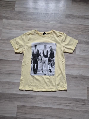 Gul t-shirt med tryck - Snygg gul t-shirt med ett stort svartvitt tryck framtill som visar tre personer bakifrån. T-shirten har rund hals och korta ärmar. Perfekt för dig som gillar streetstyle och vill ha en cool look.