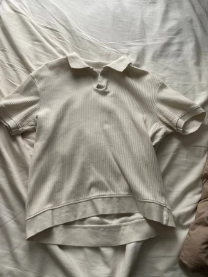 Vit ribbad pikétröja från Zara - Säljer en stilren vit pikétröja från Zara i ribbad bomull. Tröjan har korta ärmar, klassisk krage och knappslå framtill. Perfekt för en clean och avslappnad look.