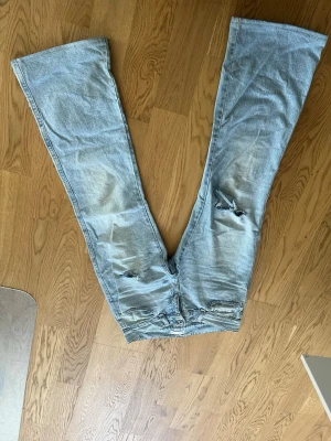 Ljusblå slitna bootcut jeans - Unika ljusblå jeans med bootcut passform och slitningar på framsidan. Baksidan har två olika färgade bakfickor, en svart och en vit, vilket ger en cool kontrast. Tillverkade i denim med klassisk knappgylf. Perfekta för dig som vill sticka ut.