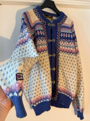 Mönstrad norsk lusekofta från Nordstrikk - Stickad kofta i klassisk norsk stil från Nordstrikk. Färgerna är blå, vit, rosa och lila med traditionellt mönster och metallspännen framtill. Rund halsringning och ribbade muddar. Norsk flagga på ärmen. Perfekt för dig som gillar retrovibbar och unik design.  Mindre fläck och skadar spänne kan ses på de sista bilderna. Inget märkvärdigt enligt mig och går att laga för de kunniga! 