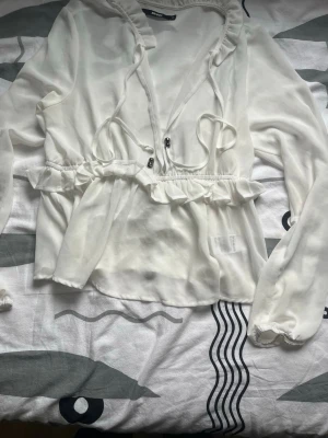 Vit blus med volang från Bik Bok - Söt vit blus från Bik Bok i tunn, luftig polyester. Blusen har långa ärmar, volangdetaljer vid midjan och knytband framtill. Perfekt för en trendig och feminin look. Passar till både jeans och kjol.