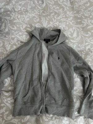 Grå hoodie från Polo Ralph Lauren - Klassisk grå hoodie från Polo Ralph Lauren med dragkedja framtill, två fickor och broderad logga på bröstet. Barnstorlek L men sitter som S 