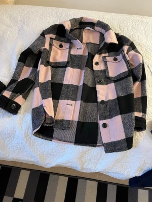 Rutig overshirt i rosa och svart - Overshirt i tjockt tyg med stora rutor i rosa, svart och grått. Två bröstfickor med lock och knappar, samt knappar längs hela framsidan och vid ärmsluten. Avslappnad passform och klassisk krage. Perfekt för lager på lager.