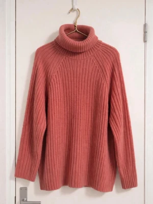 Rosa ribbstickad polotröja - Mysig rosa ribbstickad polotröja med hög krage och långa ärmar. Tröjan har en avslappnad passform och är tillverkad i ett mjukt, ull-liknande material som känns skönt mot huden. Perfekt för kyliga dagar och enkel att styla med jeans eller kjol.