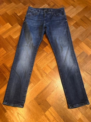 Blå raka/slim jeans från Jack & Jones  - Säljer ett par klassiska blå jeans från Jack & Jones  med rak passform och fem fickor. Jeansen har snygga slitningar och kontrastsömmar, tillverkade i denim med en autentisk tvätt. Perfekta för en avslappnad och stilren look.