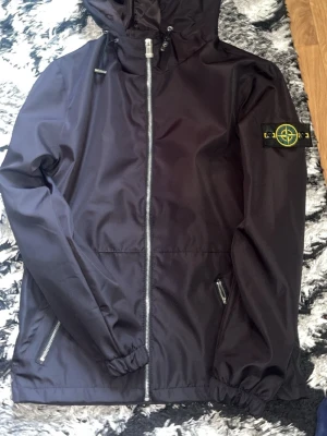Svart vindjacka från Stone Island - Snygg svart vindjacka från Stone Island med huva och dragkedja framtill. Jackan har två fickor med dragkedja och den klassiska Stone Island-loggan på vänster ärm. Materialet är lätt och vattenavvisande, perfekt för blåsiga dagar.