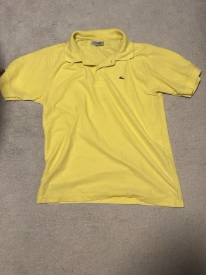 Gul pikétröja från Lacoste - Snygg gul pikétröja från Lacoste med klassisk krage och liten broderad krokodil på bröstet. Tillverkad i bomull som känns skön mot huden. Perfekt för dig som vill ha en clean och sportig look. Men känns som S