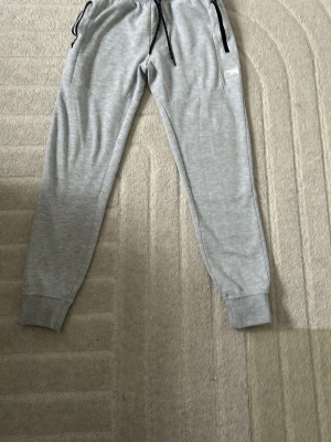 Grå mjukisbyxor med svarta detaljer - Snygga ljusgrå mjukisbyxor med svarta dragsnören och muddar vid bensluten. Byxorna har fickor med svarta dragkedjor och en liten vit logga på ena sidan. Perfekta för chill dagar eller när du vill vara bekväm.