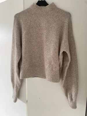 Beige stickad tröja  - Mysig beige stickad tröja från & Other Stories Los Angeles Atelier. Tröjan har hög krage, långa ärmar och ribbade muddar vid hals, ärmslut och nederkant. 