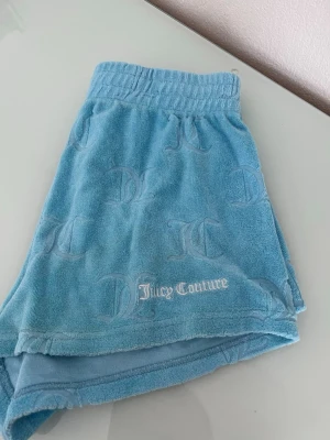 Blå frottéshorts Juicy Couture - Säljer ett par ljusblå shorts från Juicy Couture i mjuk frotté med JC-logga i relief och broderad Juicy Couture-text framtill. Shortsen har elastisk midja och avslappnad passform, perfekta för sommardagar.