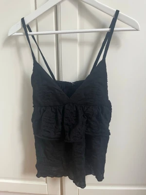 Svart volang linnetopp - Snygg svart linnetopp med tunna axelband och volangdetaljer. Toppen har v-ringning och ärmlös design, perfekt för en trendig och avslappnad look. Passar till både jeans och kjol för en cool stil.