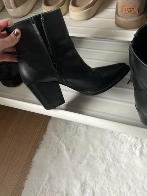 Svarta stövletter med klack - Snygga svarta stövletter i skinnimitation med spetsig tå och bred klack. Dragkedja på insidan för enkel på- och avtagning. Perfekta till höstens outfits och ger en edgy vibe till din look. Nästan som nya och ny pris är 700kr