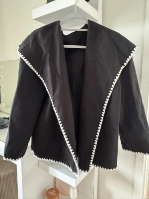 Svart kappa - Stilren svart jacka med stor krage och dekorativ vit kant runt hela jackan. Jackan har en loose passform och är tillverkad i ett mjukt tyg. Använts 1 gång säljes då jag råkat köpa den i fel storlek. Nyskick. 