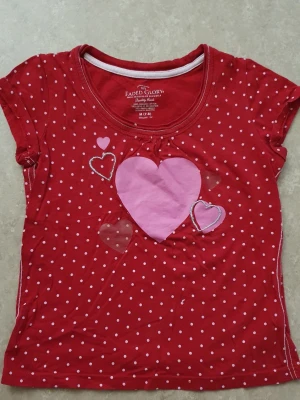 T-shirt - Söt röd t-shirt från Faded Glory med vita prickar och flera hjärtan i rosa och rött framtill. Rund halsringning och korta ärmar. Perfekt för dig som gillar färgglada och lekfulla plagg.