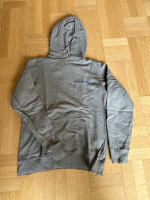 Grå hoodie från Calvin Klein Jeans - Säljer en stilren grå hoodie från Calvin Klein Jeans med liten logga broderad på bröstet. Hoodien har huva med dragsko, magficka och ribbade muddar vid ärmar och nederkant. Perfekt för en avslappnad och trendig look.