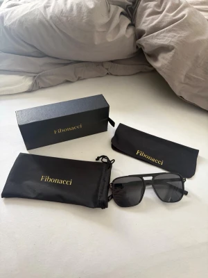Svarta solglasögon från Fibonacci - Snygga svarta solglasögon från Fibonacci med klassisk aviatorform. Glasögonen har mörka linser och metallbågar. Kommer med originalfodral och putsduk, allt i svart med guldfärgad logga.