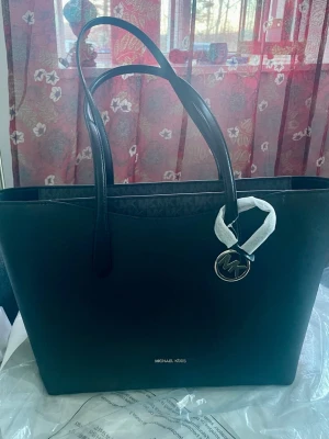*NY*Michael Kors Arden Tote bag Large - Ny med prislapp, oanvänd. Kommer i MK plastförpackning.  Svart väska med snygga grå logotypdetaljer upptill, rymmer en laptop mm.  Ord pris: 4200 sek Outlet pris: 2800 sek  Pris nu: 950 sek. Michael Kors Arden Tote kombinerar en strukturerad silhuett med generös kapacitet för vardagsbruk. Dess rymliga huvudfack, dragkedja upptill och långa axelremmar gör den idealisk för arbete, shopping och dagliga rutiner. Viktiga egenskaper: Svart | | Belagd canvas | Dragkedja upptill 