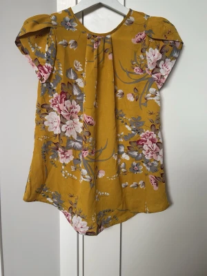 Gul blommig blus med kort ärm - Snygg gul blus med blommigt mönster i rosa, vitt och grått. Blusen har korta ärmar och en lös passform, perfekt för en fräsch och somrig look. Tillverkad i ett mjukt och lätt material som känns skönt mot huden.