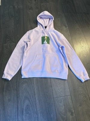 Ljuslila hoodie med tryck framtill - Säljer en ljuslila hoodie med huva och magficka. På bröstet finns ett grönt tryck med motiv av en person och ett träd. Hoodien har ribbade muddar vid ärmslut och nederkant. Perfekt för dig som gillar streetwear och vill ha något unikt i garderoben.